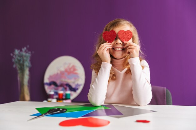 Cute little girl making a heart from paper at the table