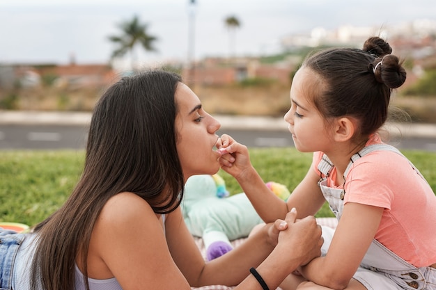 Cute little girl making up her big sister with red lipstick at city park outdoor