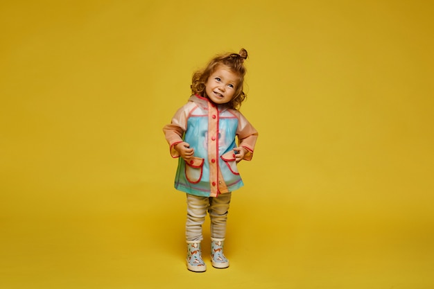 Cute little girl in a modish raincoat and rubber boots isolated at the yellow background. child fashion. copy space