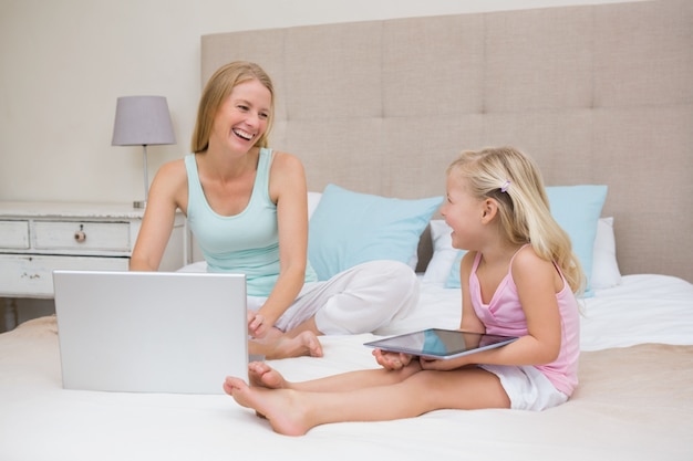 Cute little girl and mother on bed using tablet and laptop