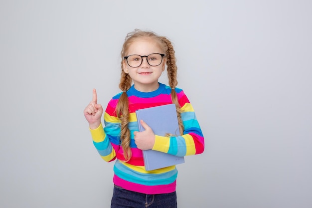 A cute little girl in a multicolored sweater and glasses holds a book on a white background smiling