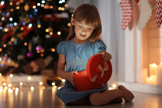 Cute little girl opening gift box in room decorated for Christmas