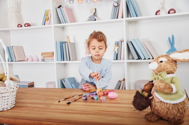 Photo cute little girl paints eggs when celebrating easter holidays indoors