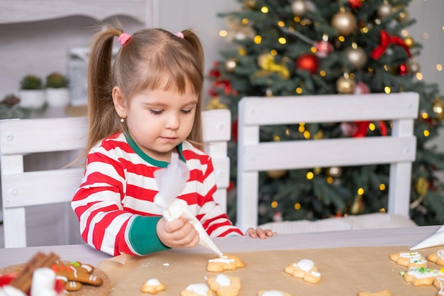パジャマ姿のかわいい女の子が、クリスマスの装飾が施されたキッチンでお祝いのジンジャーブレッドを調理