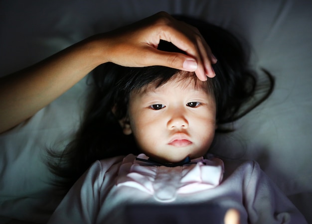 Cute little girl in pajamas playing smartphone lying on a bed with mother hand pat a head