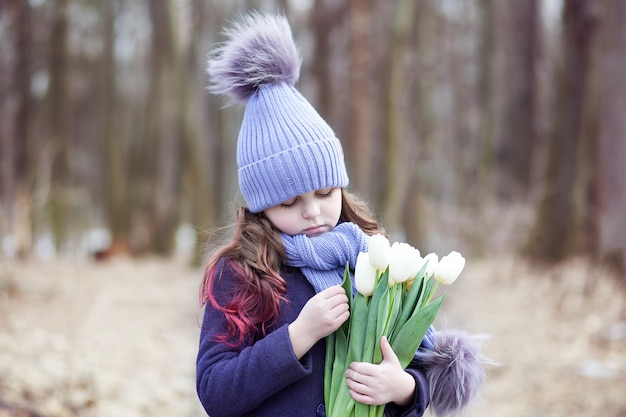 Милая маленькая девочка в парке с букетом белых тюльпанов. Цветы в подарок женскому дню матери. 8 марта. Пасха. Девушка с букетом на счастливый день матерей. Делает подарок для твоей мамы.