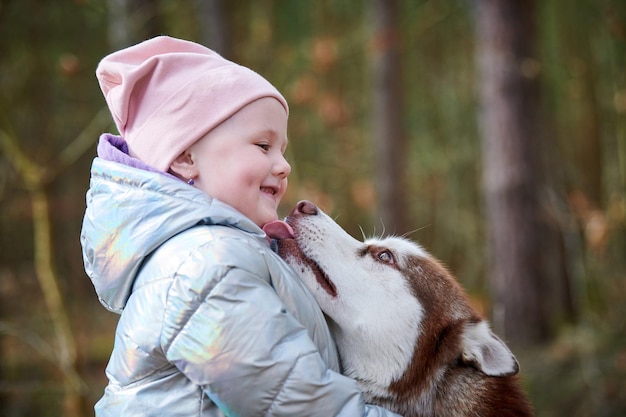 Cute little girl in pink hat and light blue jacket hugs Siberian Husky dog and smiling