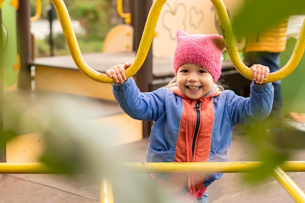 분홍색 모자를 쓴 귀여운 소녀가 도시 공원의 현대적인 놀이터에서 즐겁게 노는 카메라를 바라보고 있습니다.