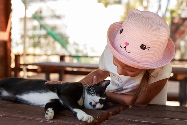 Cute little girl in pink hat petting a black cat outdoors