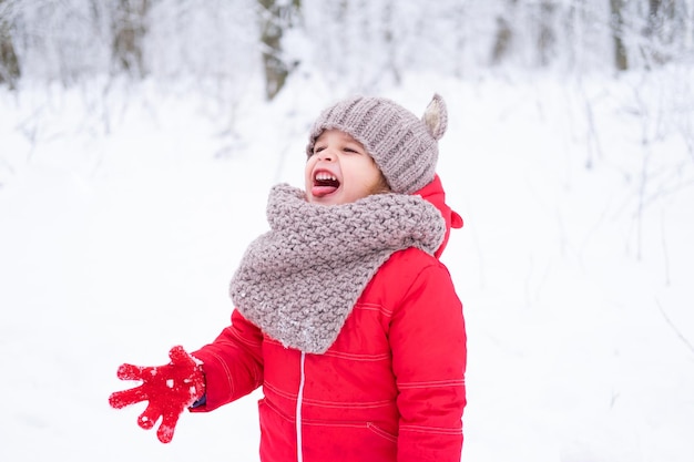 Cute little girl in pink snowsuit and knitted hat and scarf plays with snow in winter forest