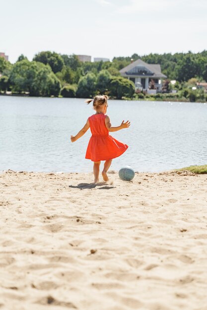 Foto bambina sveglia che gioca con la palla sulla spiaggia, sport estivi per bambini all'aperto.