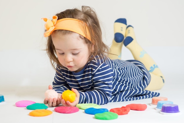 Милая маленькая девочка играет с цветным пластилином на белом фоне