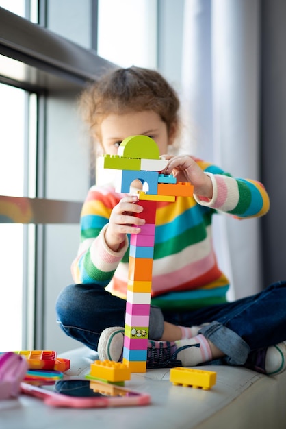 A cute little girl plays construction set. Childhood. Development. She sits near a window with a large teddy bear. The girl is dressed in a colored sweater and jeans.