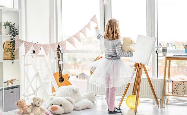 Photo cute little girl pointing at window for teddy bear in playroom