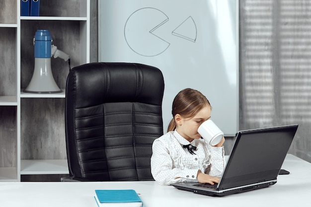 Милая маленькая девочка изображает главу агентства за белым столом, пьющего из белой чашки