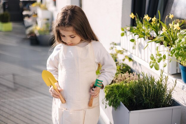 Cute little girl put a small shovel and a rake into her overall gardening on balcony