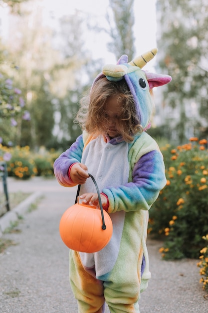 ハロウィーンの虹のユニコーンの衣装を着たかわいい女の子がカボチャのバスケットにお菓子を集めに行きます