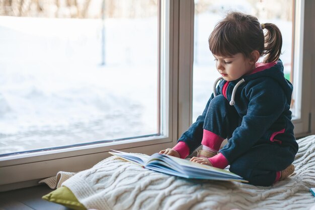 Cute little girl reading a book sitting on a woolen blanket near the window. it's winter, it's snowy and cold outside.