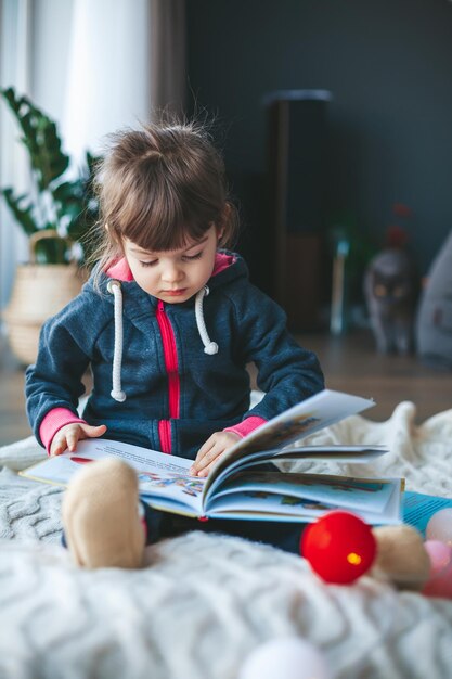 窓の近くの羊毛の毛布の上に座って本を読んでかわいい女の子。冬です。外は雪が降って寒いです。