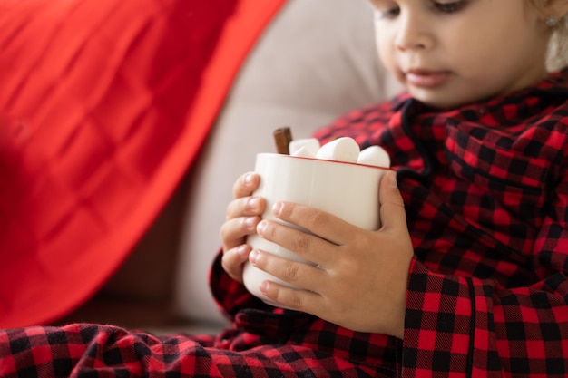 cute little girl in red Christmas pajama holding cup of cacao with marshmallows sitting on couch