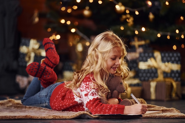 Милая маленькая девочка в красном праздничном свитере, лежа с плюшевым мишкой в помещении, празднует новый год.