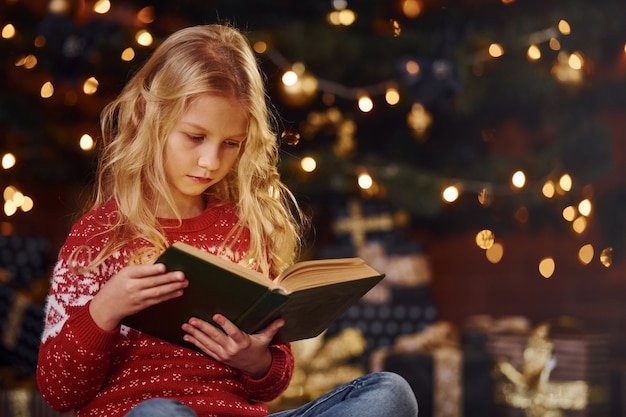 Cute little girl in red festive sweater reading book indoors at christmas holiday time.