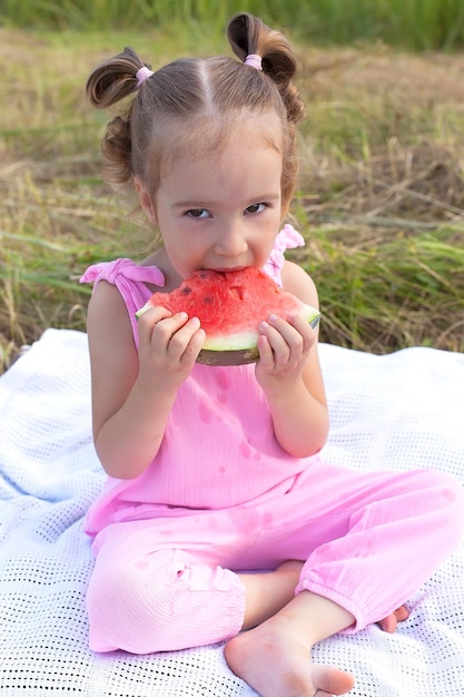 Милая маленькая девочка в красной футболке с кусочком арбуза в руках Большой кусок спелого красного арбуза в руках маленькой девочки