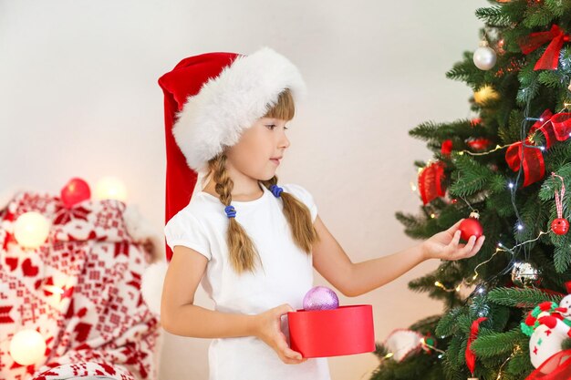 Cute little girl in Santa hat decorating Christmas tree at home