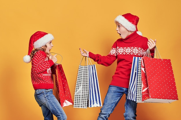 Cute little girl in Santa hat hanging shopping bag with Christmas gifts on hand of astonished boy during holiday celebration against yellow background