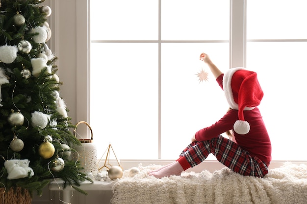 Cute little girl in Santa hat holding Christmas ornament on window sill at home