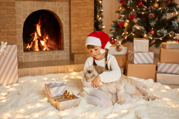 Bambina sveglia in cappello della santa che abbraccia con il cane su priorità bassa del bellissimo albero di natale