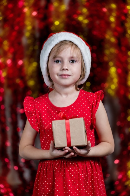 Cute Little Girl In Santa Hat is Holding Christmas gift and smiling .