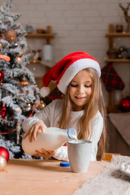 산타 모자와 잠옷을 입은 귀여운 어린 소녀가 크리스마스 모형으로 부에서 우유를 마시고 있습니다.