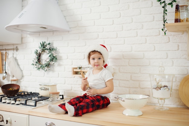 Cute little girl in santa hat, preparing cookies in the kitchen at home. Sits on the kitchen table and helps mom prepare a festive Christmas dinner	