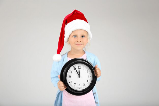 Cute little girl in Santa hat with clock on light background. Christmas countdown concept