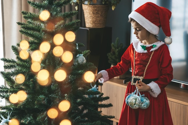 산타 의상을 입은 귀여운 소녀가 크리스마스 트리를 장식하고 휴일 추억을 만들고 있습니다.