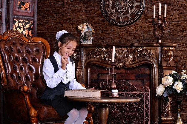 Cute little girl in school uniform reading book in room with vintage furniture