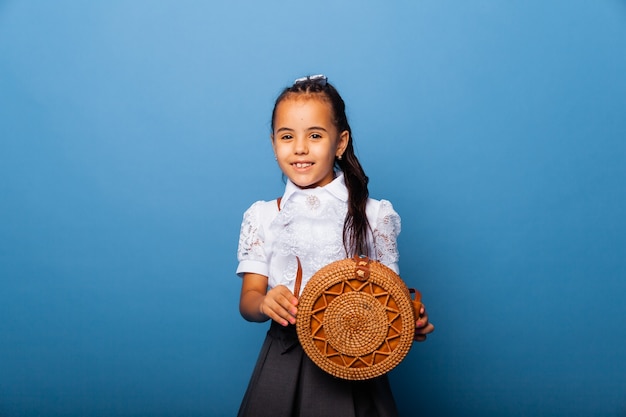 Foto carina bambina di sette anni che posa in uniforme scolastica e tiene in mano una borsa rotonda di vimini su sfondo blu