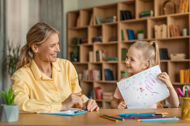 Cute little girl showing woman psychologist her drawing kid having therapy session with child development specialist