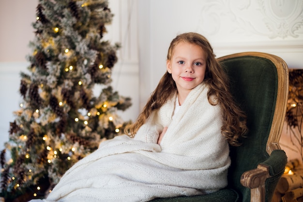 Cute little girl sits by the christmas tree wrapped in a warm blanket at home