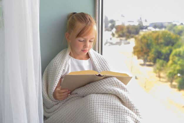 A cute little girl sits by the window and reads a book in the room at home Beautiful autumn nature