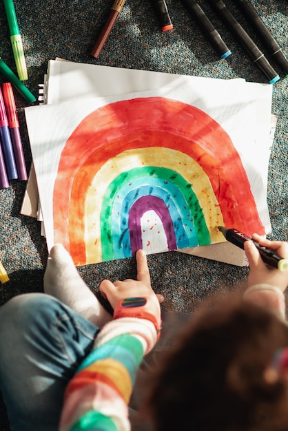 A cute little girl sits on the floor in the rays of the sun and draws a rainbow with colorful marker