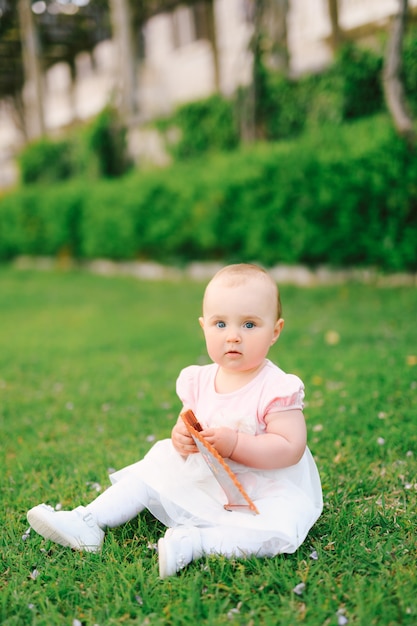 Cute little girl sits on the green grass in the garden and holds a folding fan in her hands villa