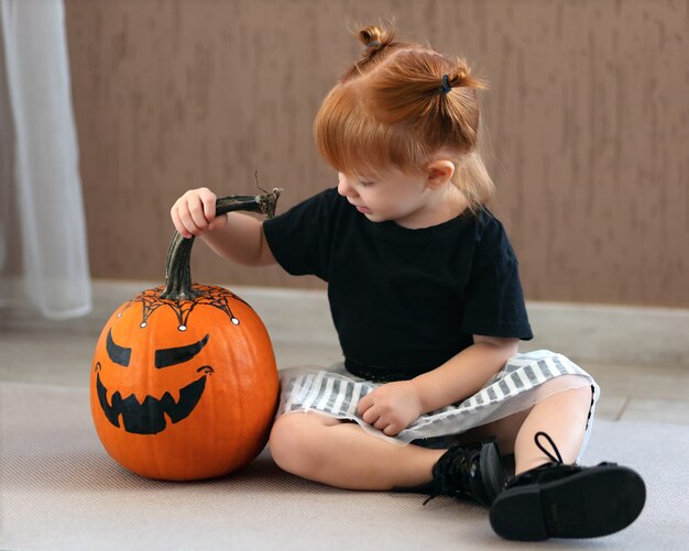 Cute little girl sits and looks at a painted pumpkin for halloween in anticipation of the holiday