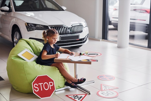 Cute little girl sits on the soft green chair by the table with pencil and paper sheets. Near modern automobile and road signs on the floor.