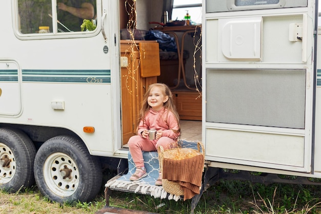 Cute little girl sits on the steps of a motorhome and drinks hot chocolate with marshmallows