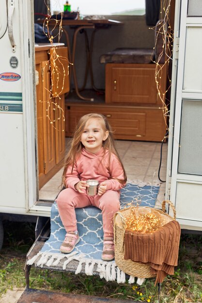 Cute little girl sits on the steps of a travel trailer and drinks hot chocolate with marshmallows