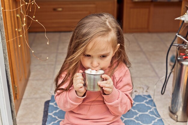 Cute little girl sits on the steps of a travel trailer and drinks hot chocolate with marshmallows