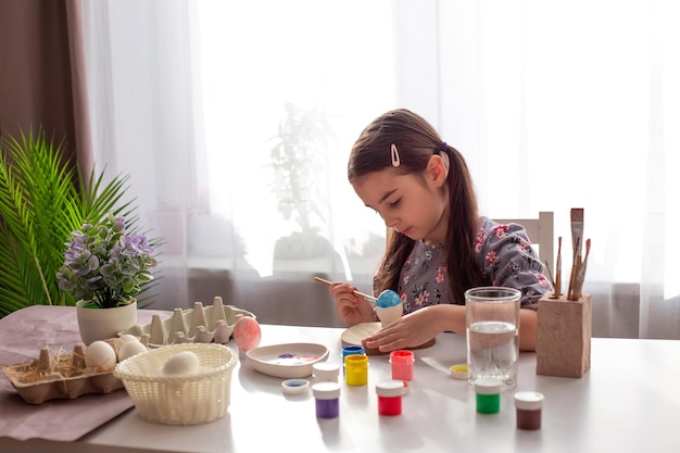 A cute little girl sits at a white table by the window paints eggs with a brush and paint in jars