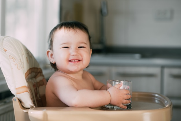 Cute little girl sitting in a baby chair in the kitchen and drinking water